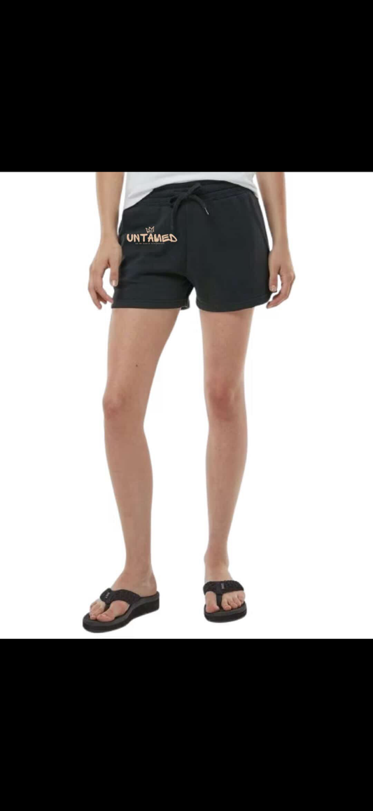 Black athletic shorts with a drawstring waist and a small gold 'UNTHRED' logo on the left hip, shown on a model from the waist down wearing black sandals against a white background.